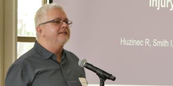 Image of Randy Huzenic, a distinguished man with glasses and a beard standing before a microphone