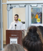 Mitch Bell speaks at a podium in front of a large crowd during the opening of the Assist+ center